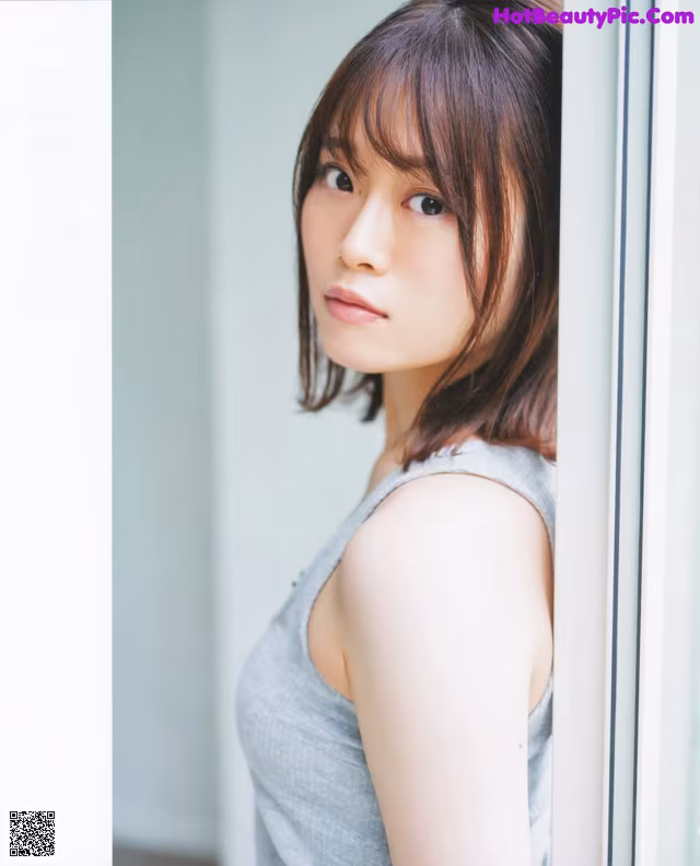A woman in a gray tank top leaning against a window.